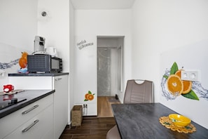 A modern kitchen with a coffee machine, a kettle, and a red piggy bank on the counter. There is a wall decal with an orange slice and a door with a welcome message.