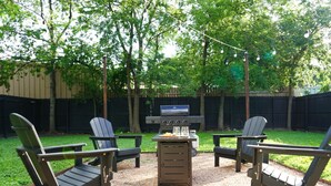 Patio trasero con una parrilla, cuatro sillas tipo Adirondack y una mesa con comida y bebidas.
