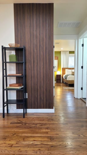 A room with a wooden floor, a black bookshelf with books and a basket, a bed with a bedside table and lamp, and a sliding door leading to another room.