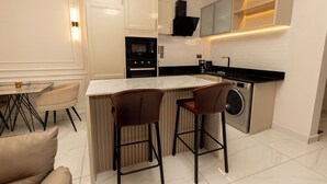 A modern kitchen with a central island, bar stools, a built-in washing machine, and a dining area with a table and chairs. - Charming 1BR Suite | Rooftop Pool | Scenic City Views (Accra)