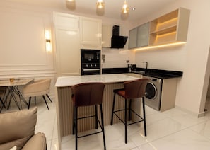 A modern kitchen with a central island, bar stools, a built-in washing machine, and a dining area with a table and chairs. - Charming 1BR Suite | Rooftop Pool | Scenic City Views (Accra)