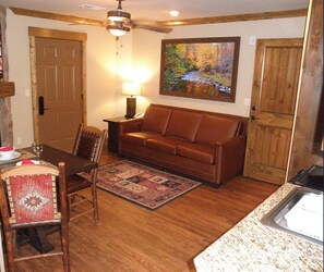 Ein Zimmer mit Holzboden, einem braunen Ledersofa, einem Esstisch mit Stühlen, einem Teppich, einem Gemälde und einem Deckenventilator.
