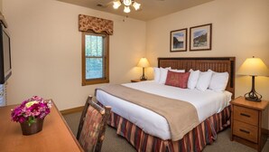 Ein Hotelzimmer mit Bett, zwei Nachttischen mit Lampen, einem Holzkopfende, einem Fenster mit Jalousien, einem Deckenventilator und einem kleinen Schreibtisch mit Blumenvase.