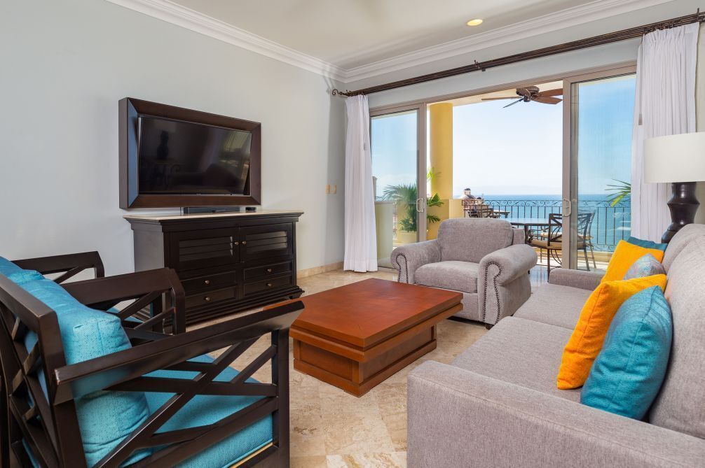 A living room with a flat-screen TV, dark wooden furniture, a grey sofa with colorful cushions, and a view of the ocean through large sliding glass doors.