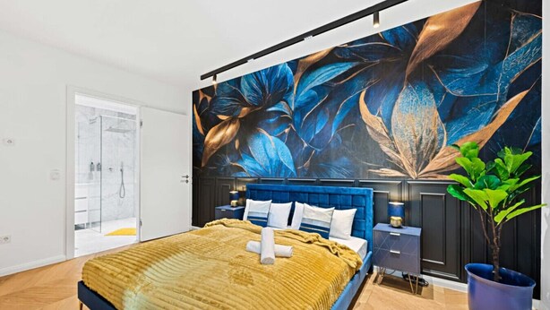 A bedroom with a large bed, a blue headboard, a nightstand, a potted plant, and a wall mural.