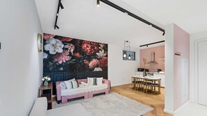 A modern living room with a large floral mural, a pink sofa, and a dining area with a white table and chairs.