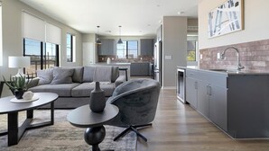 Een moderne woonkamer met een grijze bank, een ronde salontafel en een grijze fauteuil. Er is een keuken met grijze kastjes en een achterwand van baksteen.