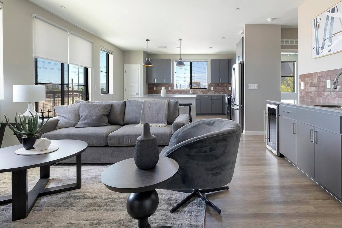 Un salon moderne avec un canapé gris, une table basse ronde et un fauteuil gris. La cuisine est dotée de meubles gris et d’un mur de fond en briques.