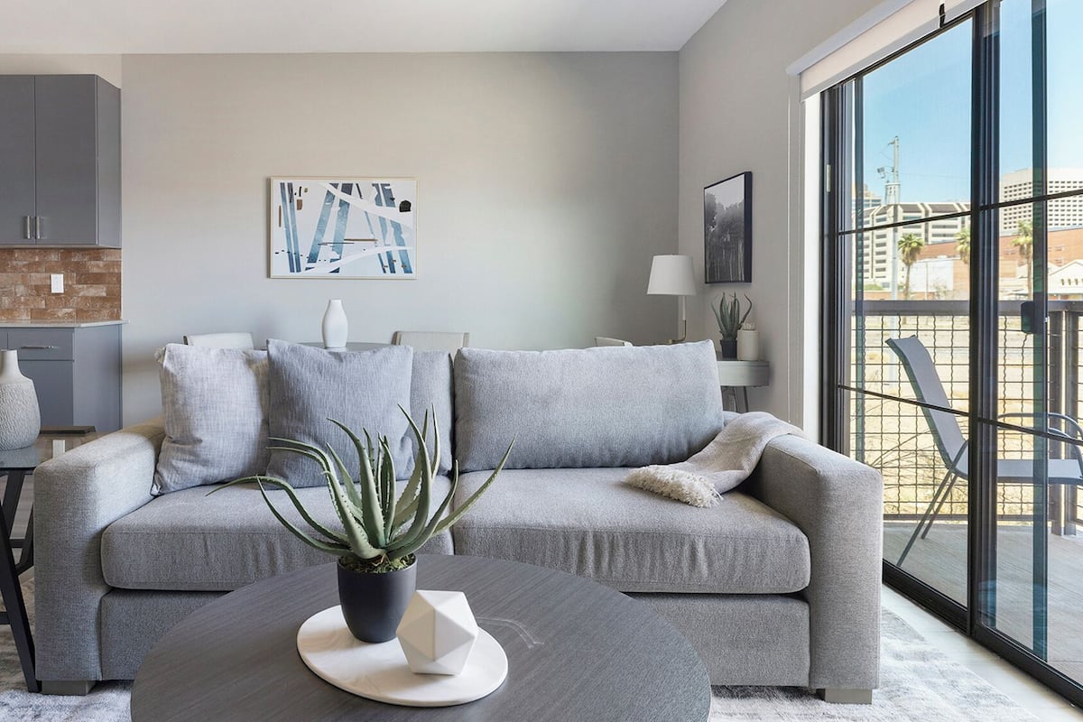 Un salon moderne avec un canapé gris, une table basse et une plante en pot. On y trouve également un coin cuisine avec des meubles gris et un mur de fond en briques. Une grande fenêtre offre une vue sur l’extérieur.