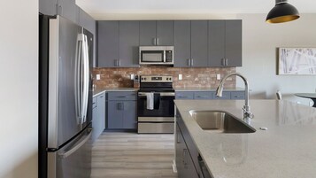 A modern kitchen with grey cabinets, a stainless steel refrigerator, a microwave, an oven, and a sink with a granite countertop.