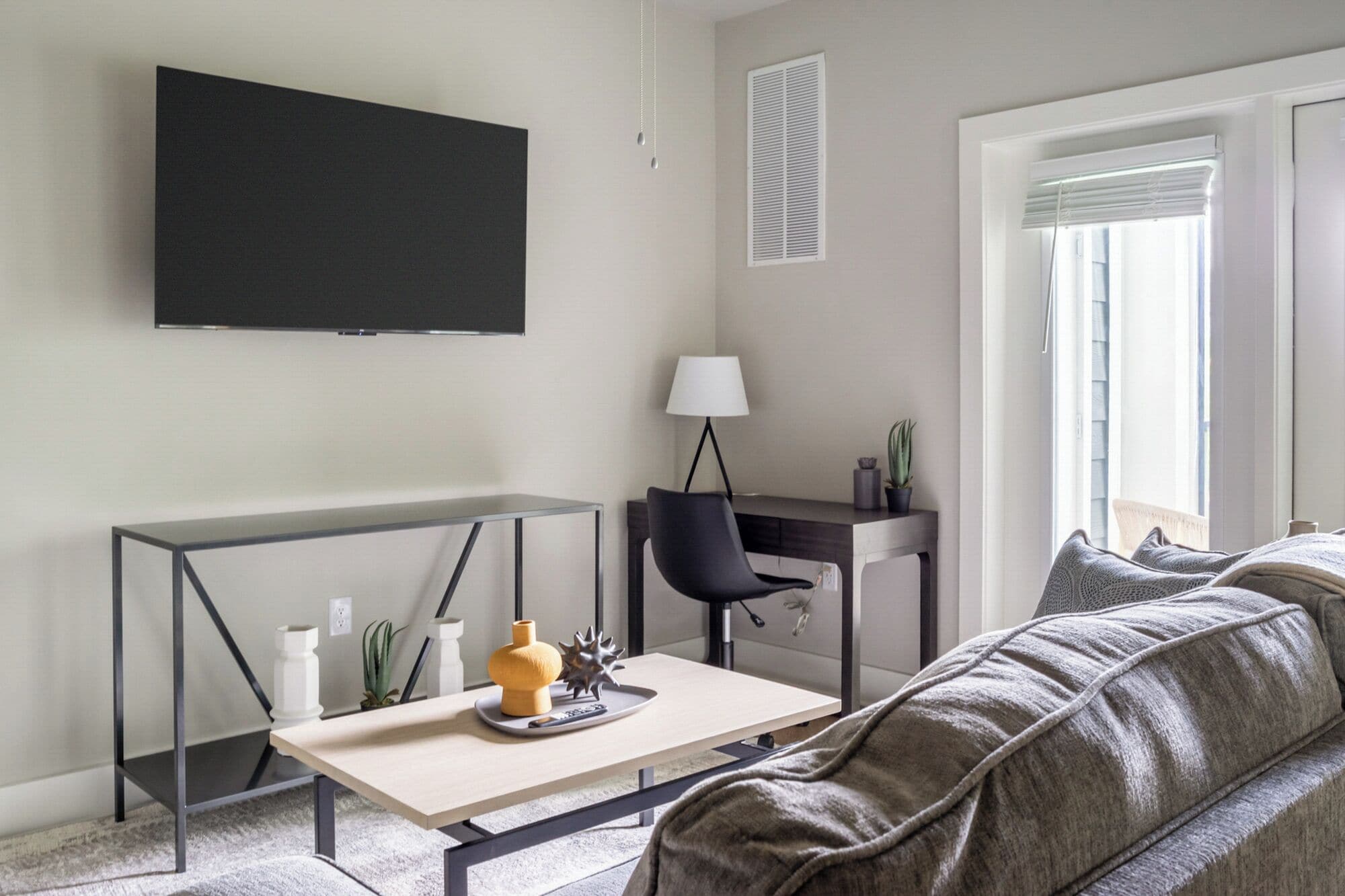 A modern living room with a flat-screen TV mounted on the wall, a desk with a chair, a sofa, and a lamp.