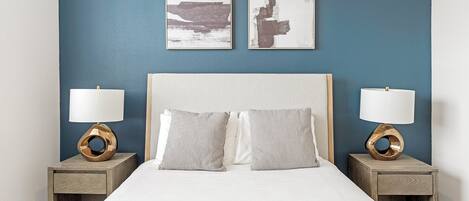 A bedroom with a bed, two bedside tables, two lamps, and two wall art pieces.