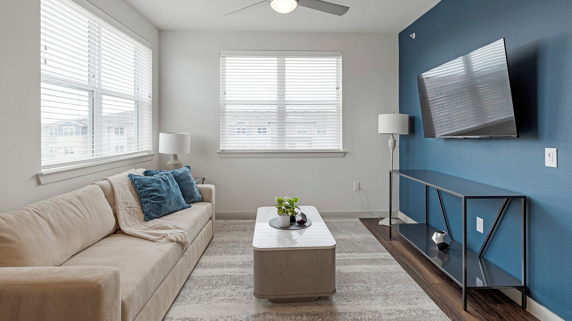 A modern living room with a beige sofa, two blue cushions, a coffee table with a plant, and a TV mounted on a blue wall.