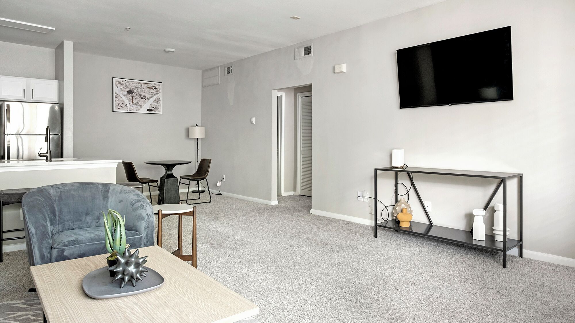 A modern living room with a grey sofa, a dining table, a TV mounted on the wall, and a small console table with decorative items.