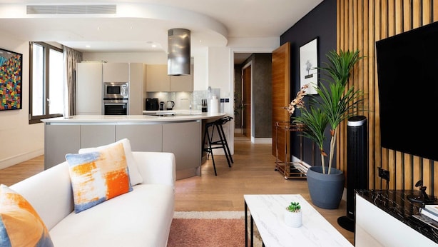 A modern living room with a white sofa, a coffee table, and a TV. There is a kitchen with a large island and a dining area with bar stools. - Luxury Apartment Near Notting Hill (London)