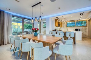 A modern dining area with a wooden table and light grey chairs, a blue vase with flowers, and a kitchen with a large TV.