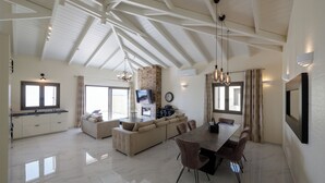 A spacious living area with a high ceiling, a fireplace, a dining table with chairs, and a large window.