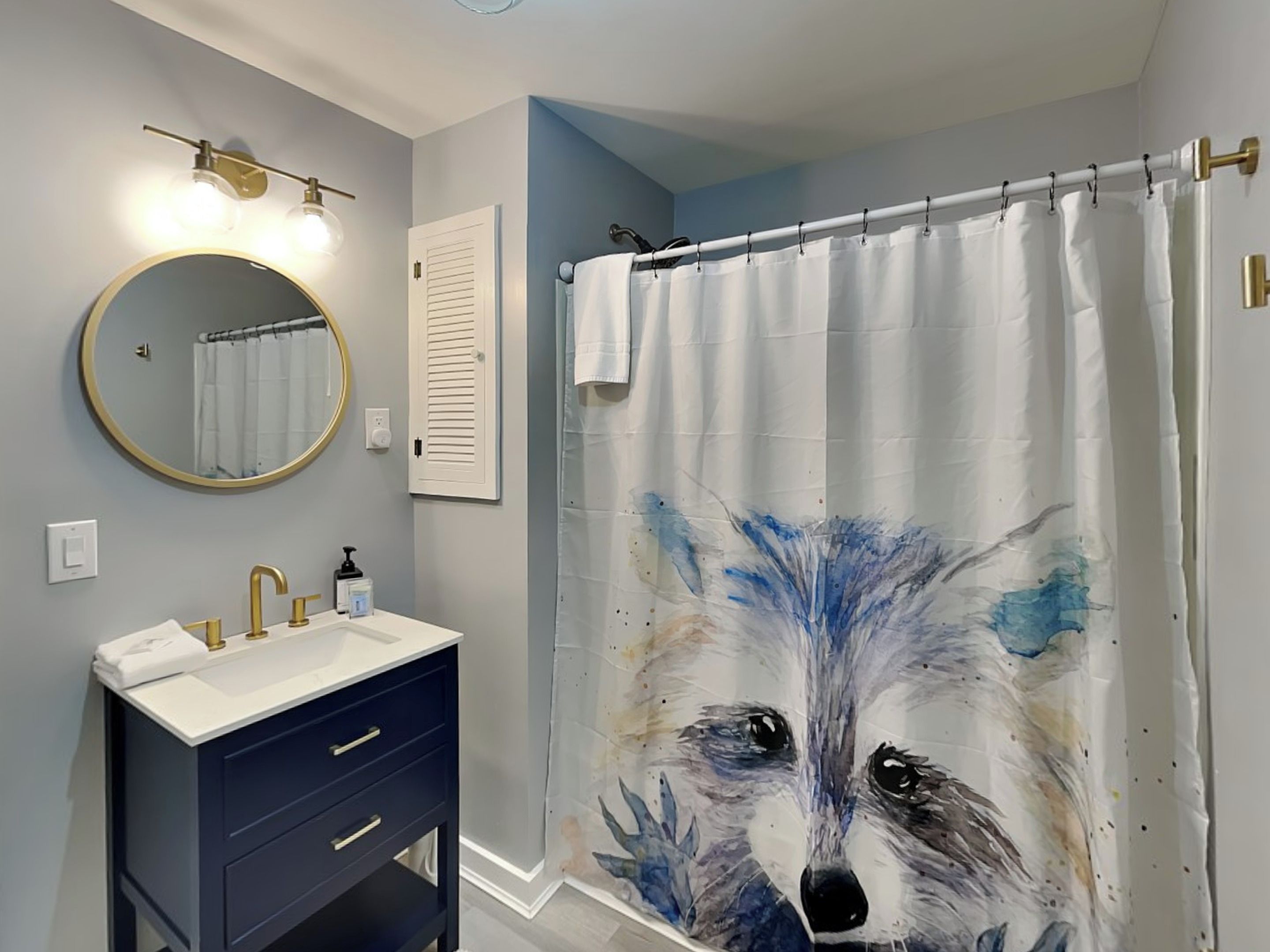 Une salle de bain avec un miroir rond, un lavabo doté d’un robinet doré, un meuble-lavabo bleu foncé et un rideau de douche orné d’un motif représentant un loup.
