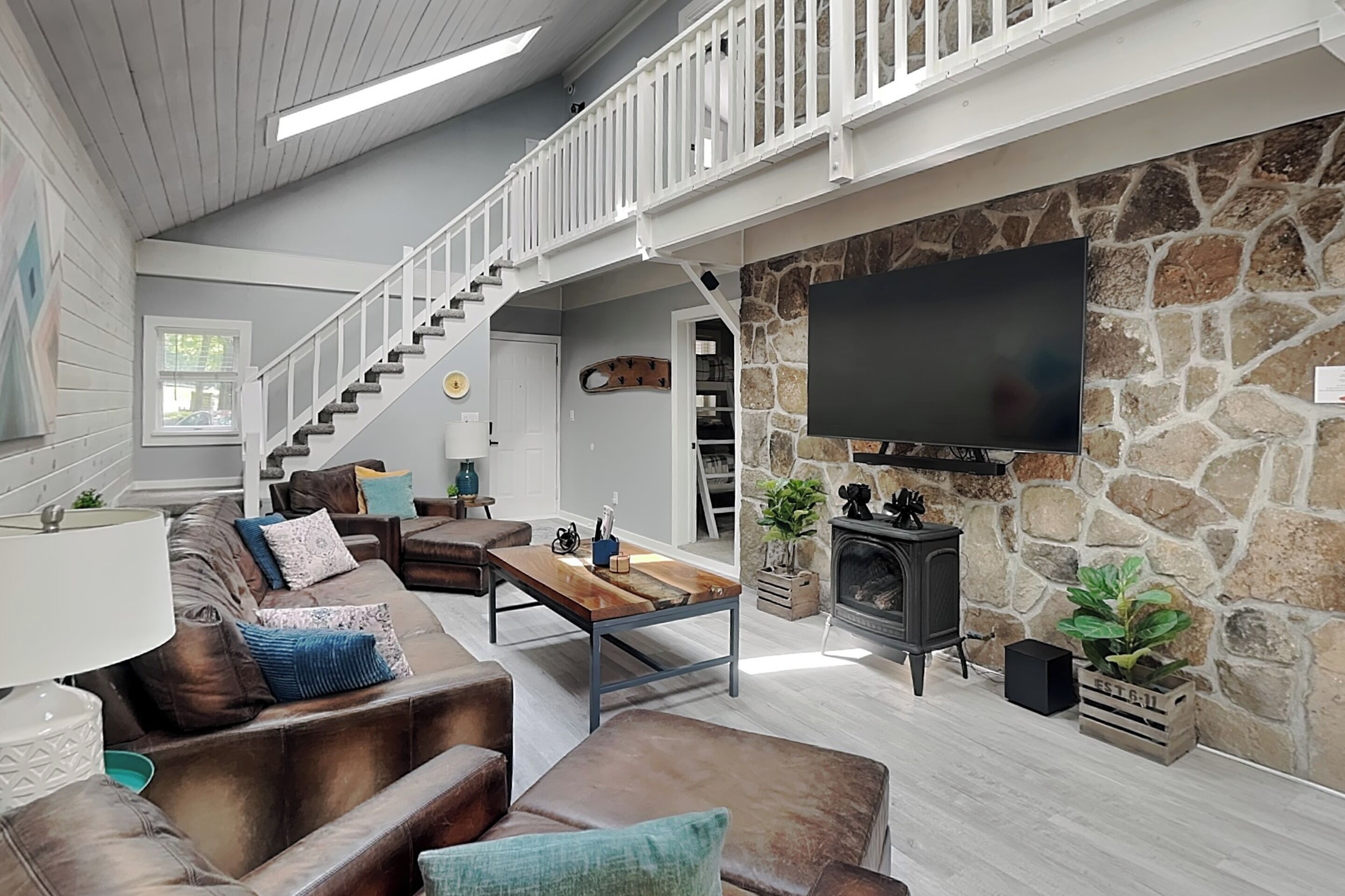 Un salon spacieux doté d’une cheminée en pierre, d’un grand téléviseur à écran plat fixé au mur, d’une table basse en bois et d’un canapé en cuir agrémenté de coussins décoratifs.