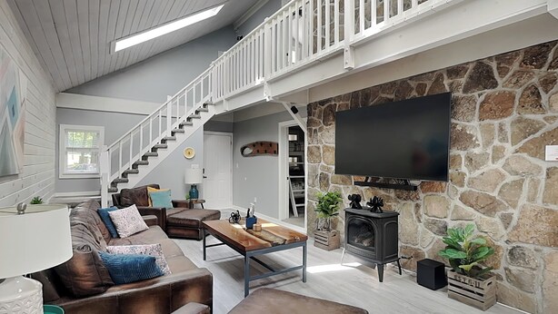 Amplia sala de estar con chimenea de piedra, un televisor de pantalla plana grande montado en la pared, una mesa de centro de madera y un sofá de cuero con cojines decorativos.