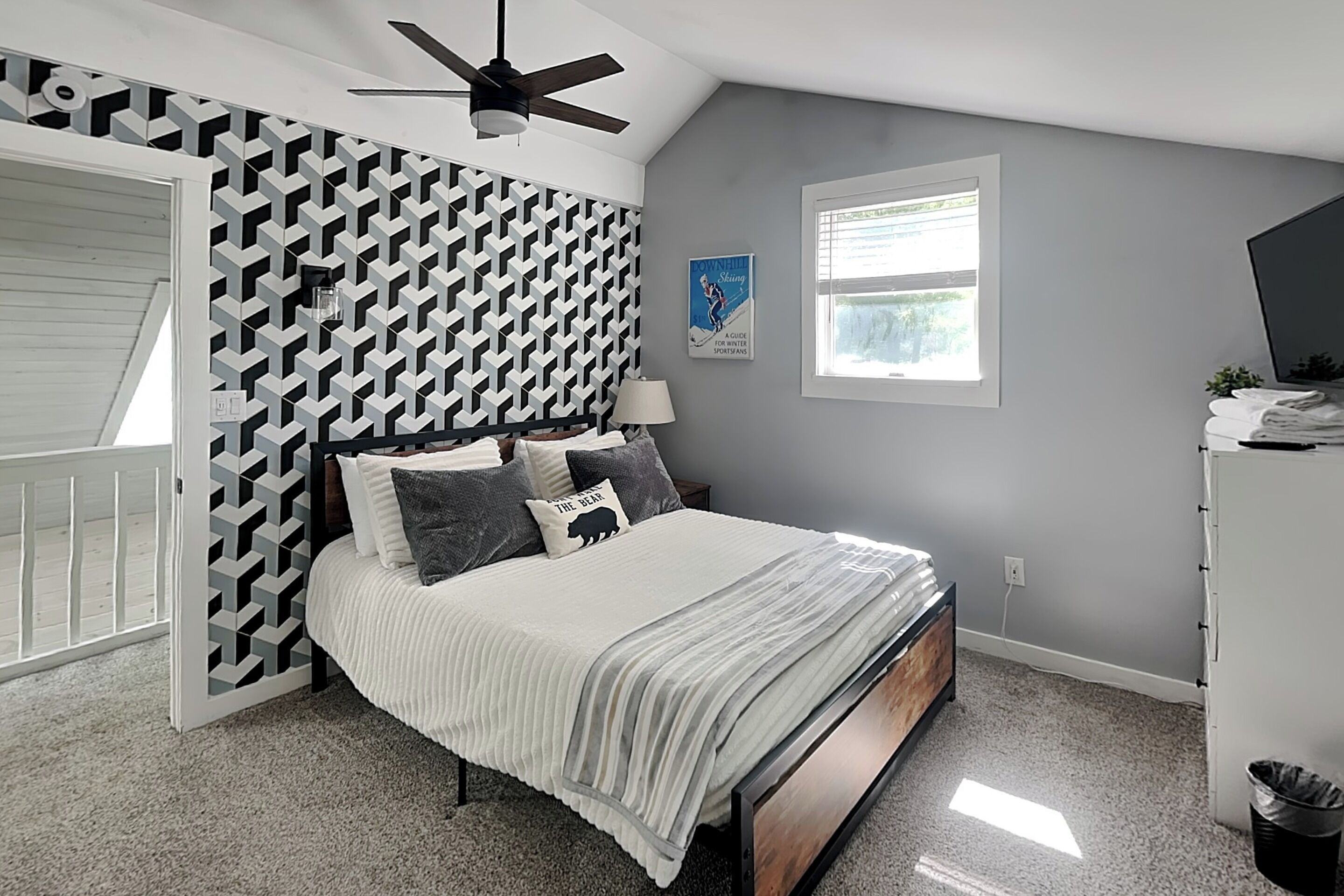A bedroom with a bed, a ceiling fan, a window, a wall with a geometric pattern, and a door.