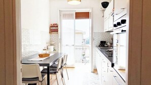 A narrow kitchen with a dining table and chairs, a small refrigerator, and a window overlooking a balcony.