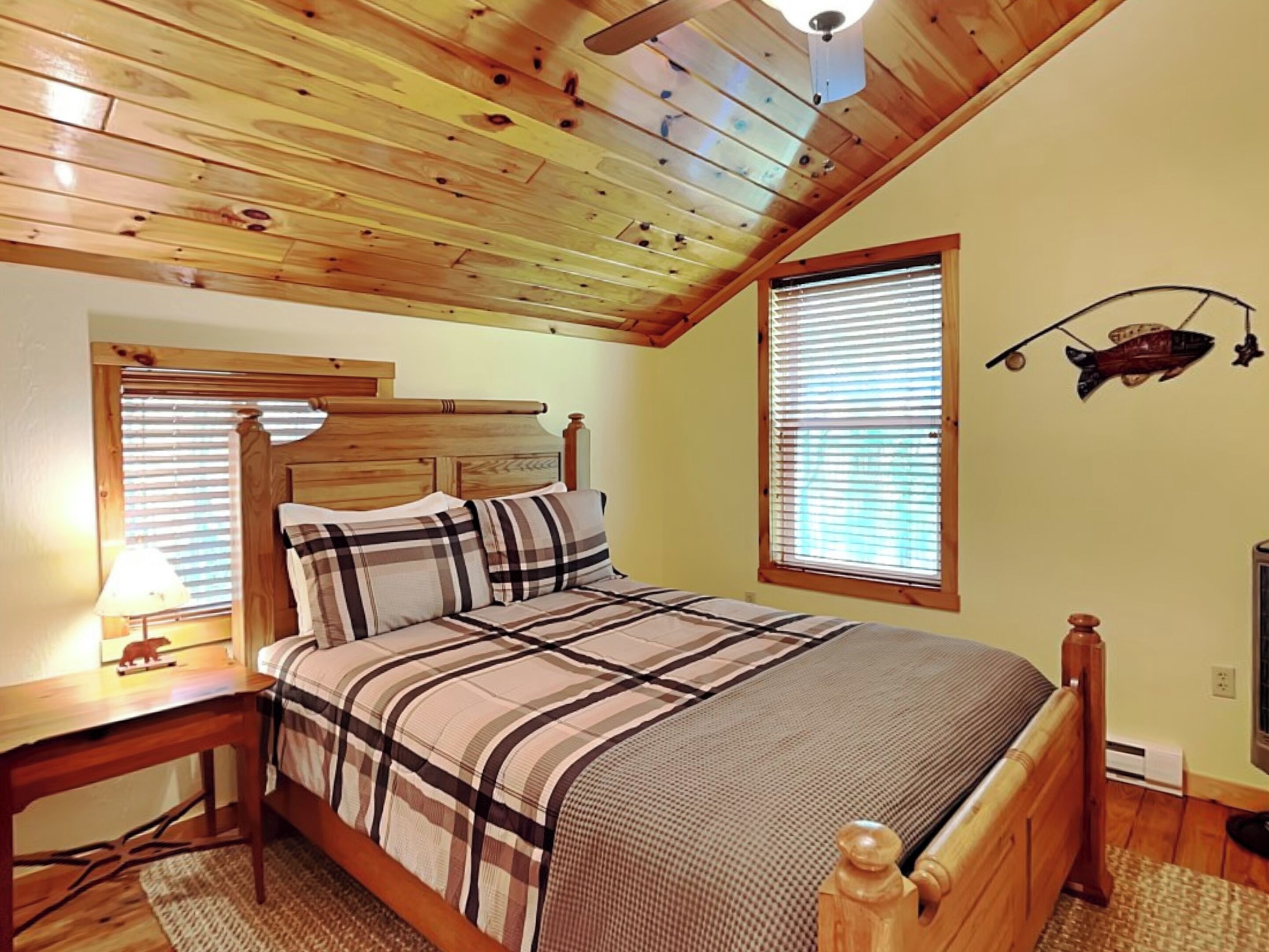 Um quarto com teto de madeira, uma cama com colcha xadrez, uma mesa de cabeceira com abajur e uma janela com persianas.