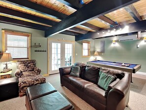 A cozy living room with a brown leather sofa, a patterned armchair, a pool table, and a ceiling with exposed beams. - Campfire Lodge by Taylor Made Deep Creek (Swanton)