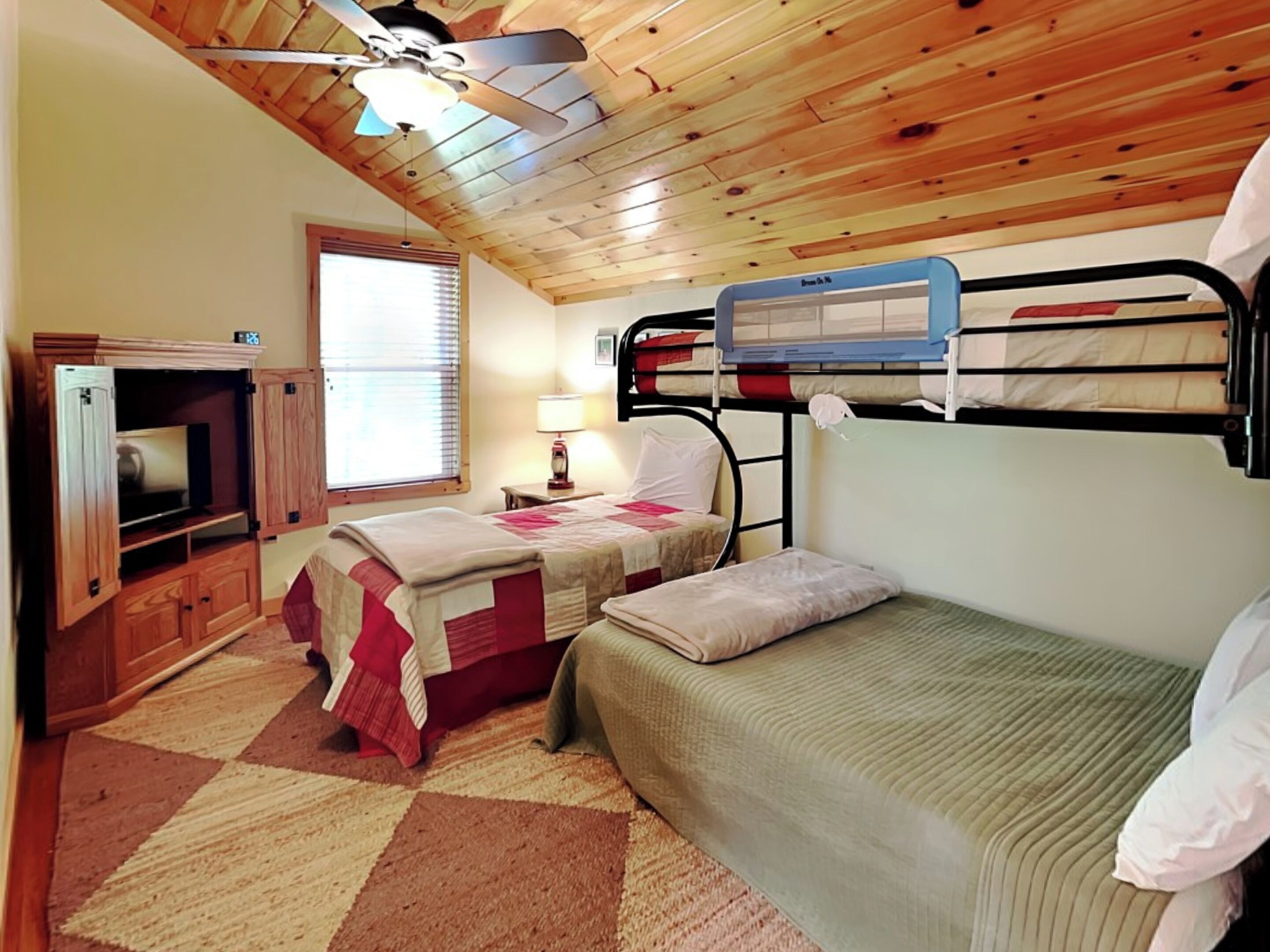 A room with wooden ceiling, bunk beds, a TV, and a window with blinds.