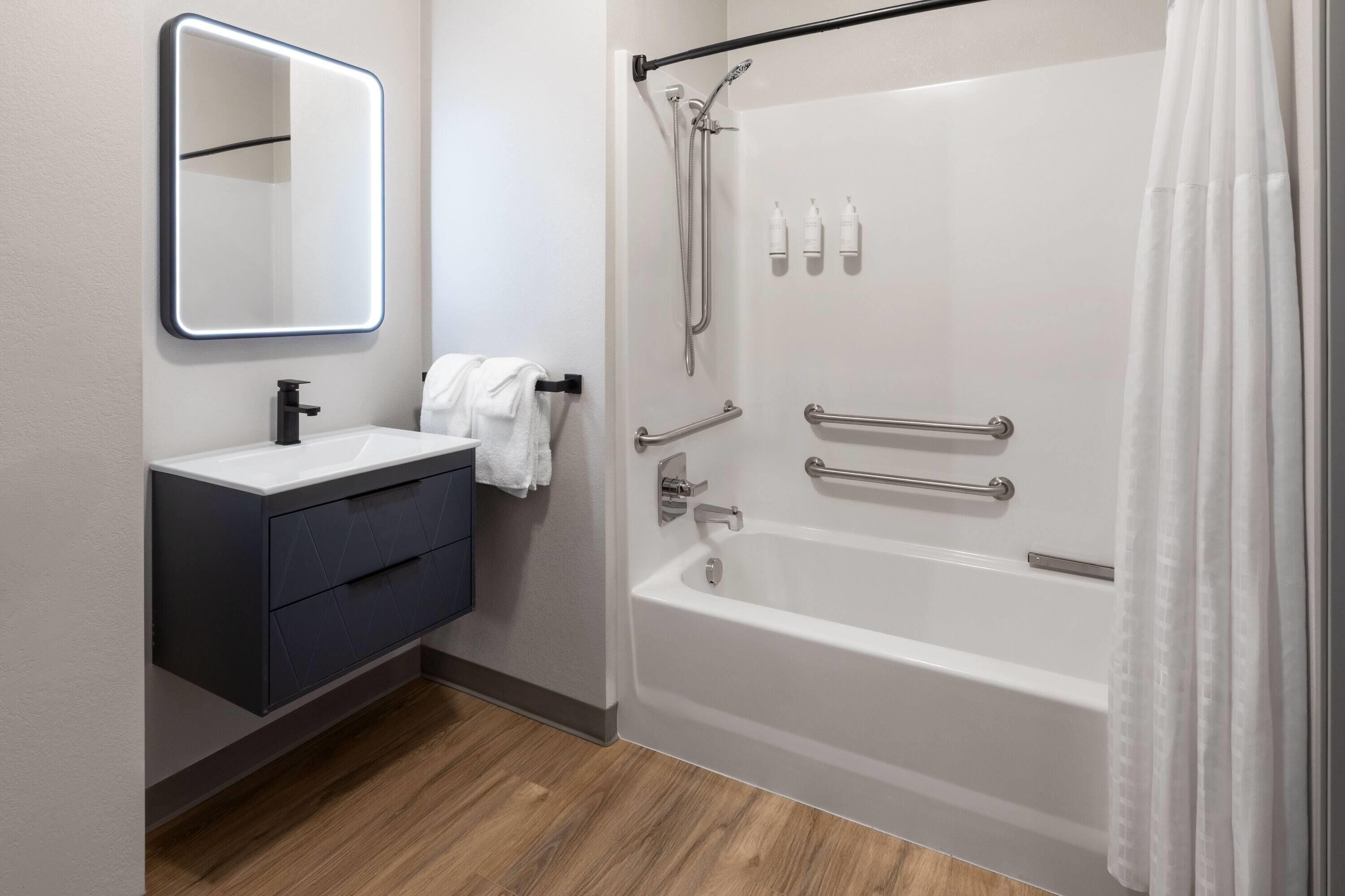 A modern bathroom with a white bathtub, a dark vanity, a mirror, and a shower with grab bars.