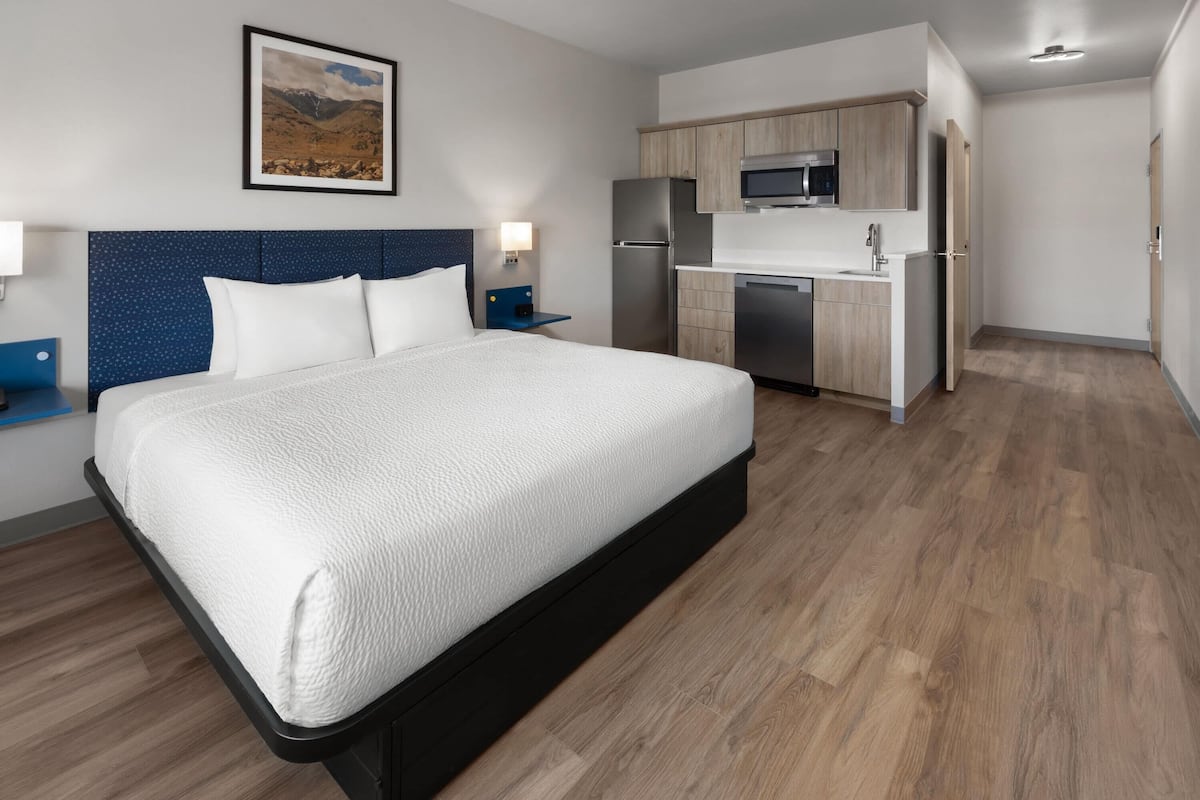 A modern hotel room with a large bed, a kitchenette, and a framed picture on the wall.