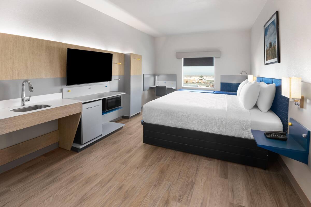 A modern hotel room with a bed, a desk with a sink, a flat-screen TV, and a window with a city view.