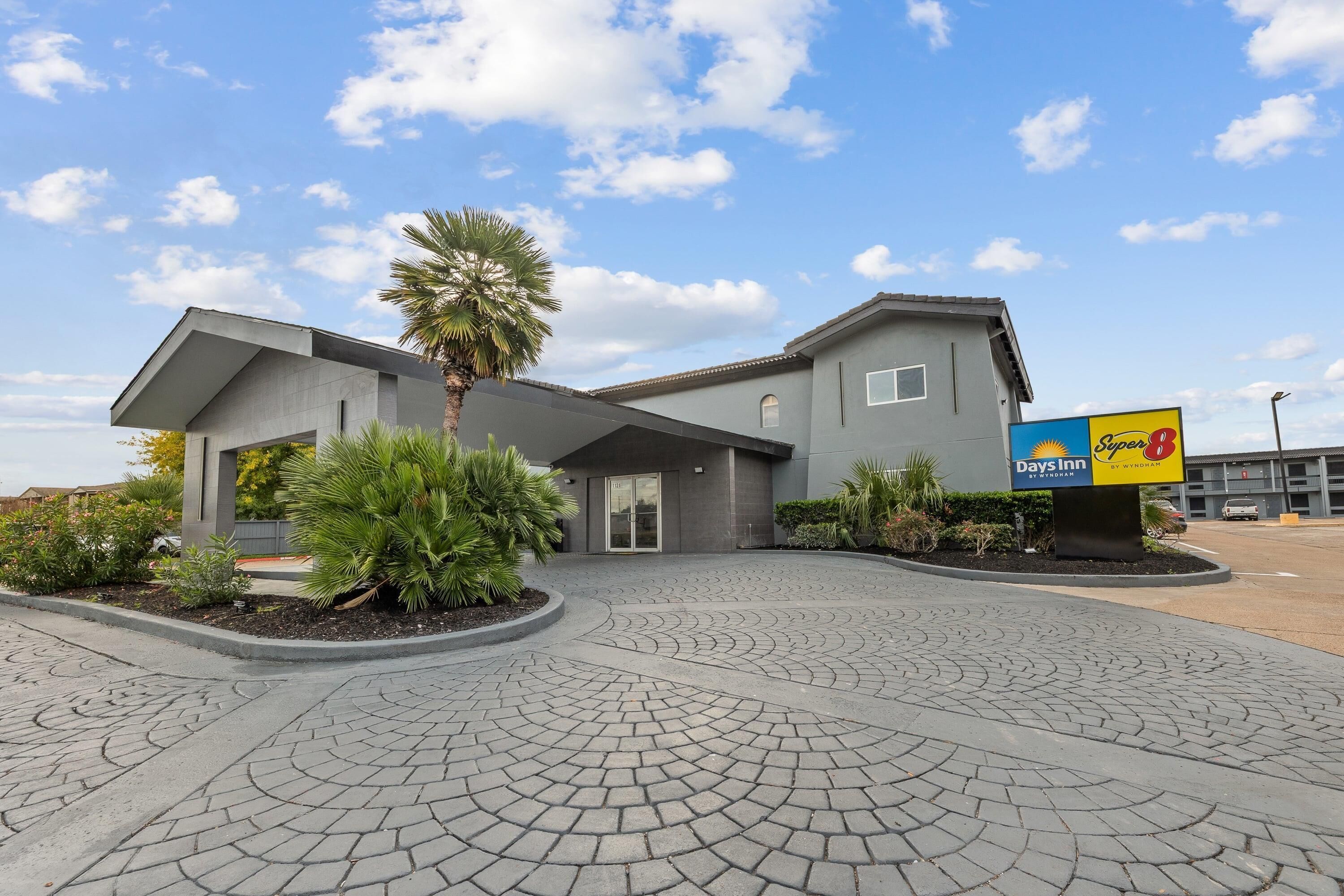 A Super 8 Days Inn with a modern design, featuring a curved driveway and landscaped areas with palm trees and shrubs.