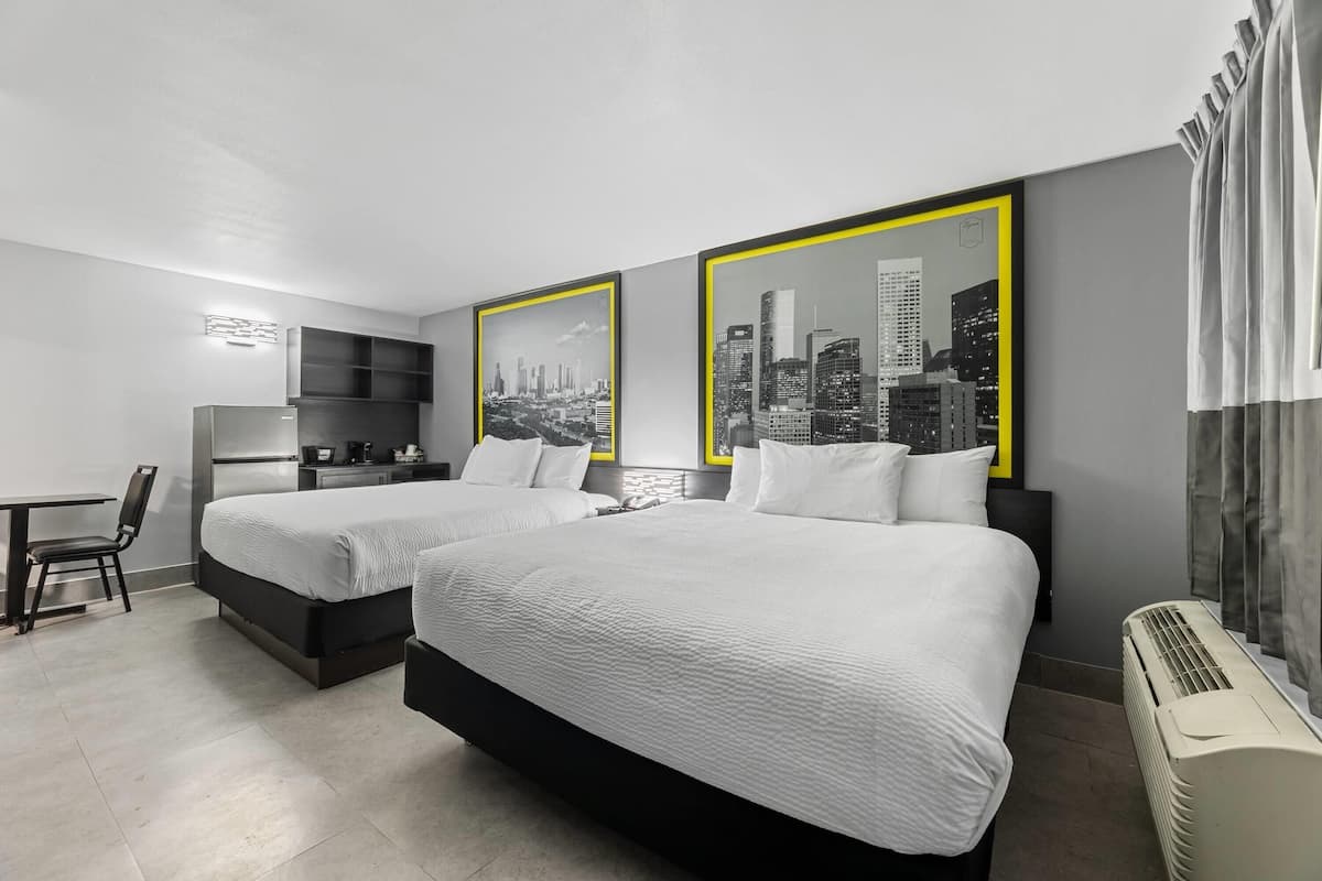 A hotel room with two beds, a desk, a chair, a refrigerator, and a heater.