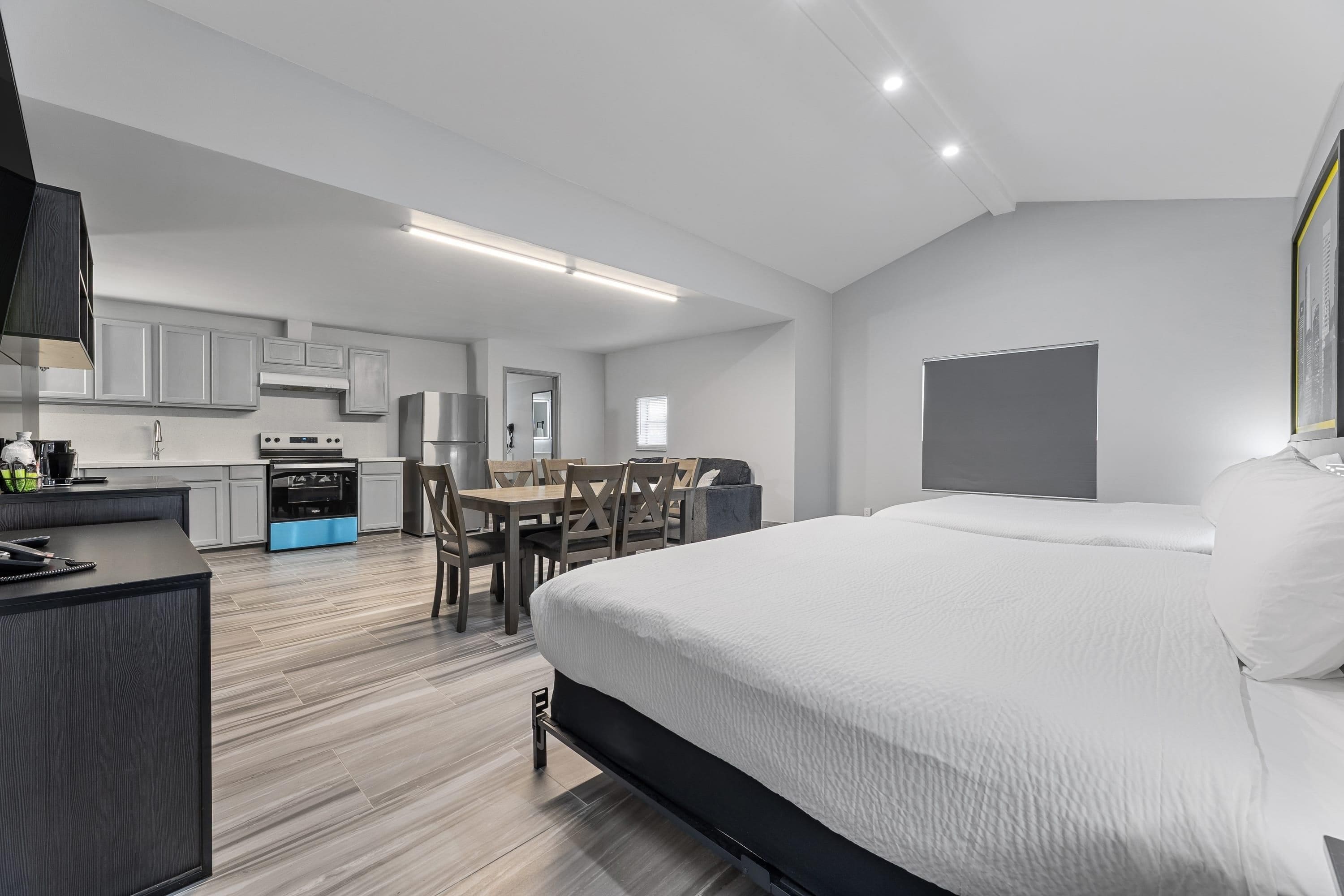 A modern hotel room with a large bed, a dining area, and a kitchenette.