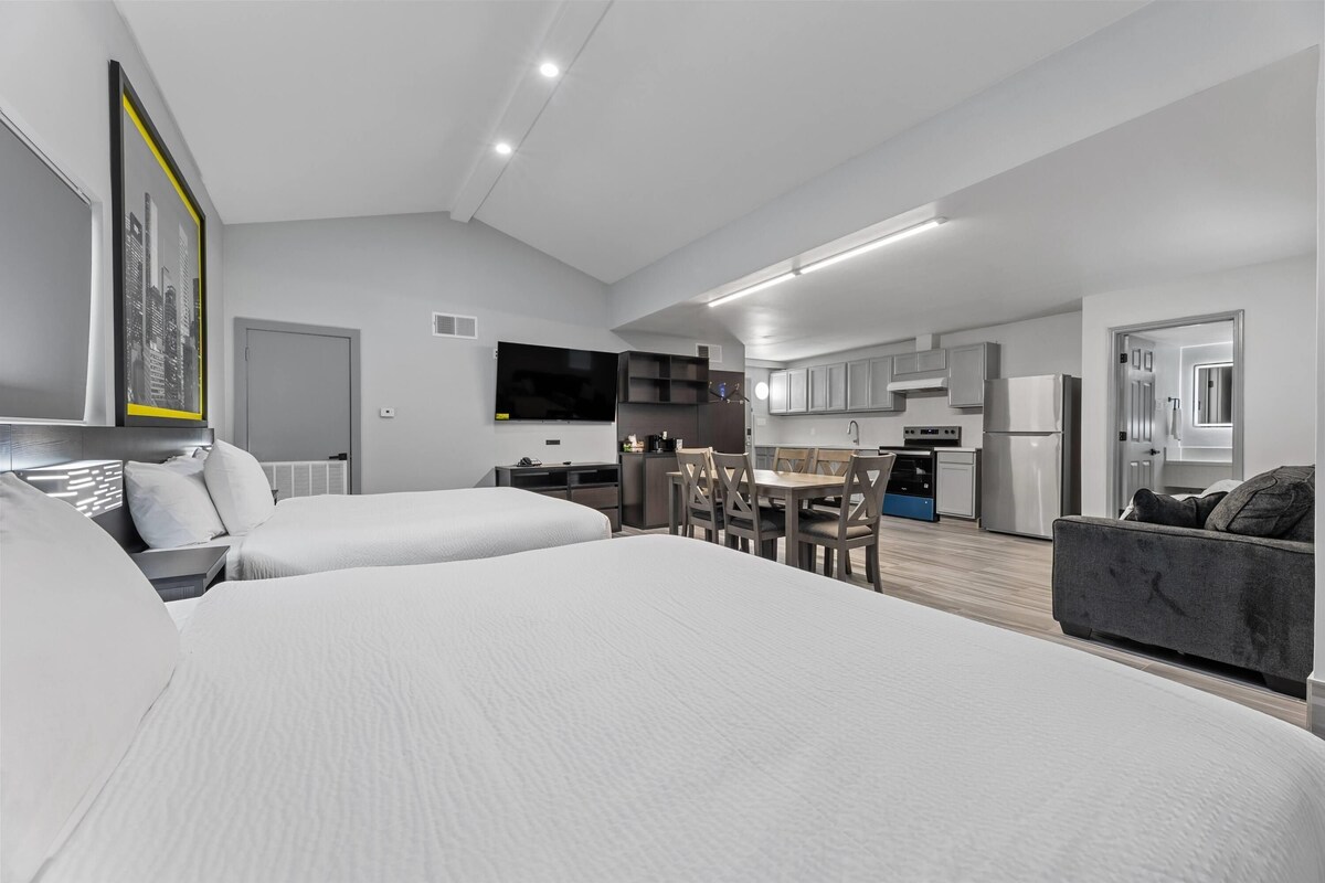 A modern hotel room with a large bed, a desk, a dining area, and a kitchenette.