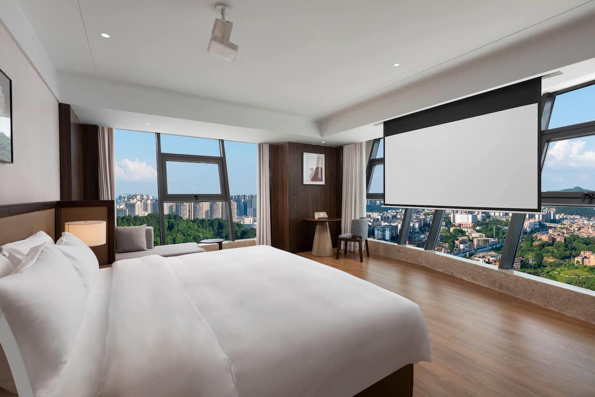 A modern hotel room with a large bed, a desk, and a view of a cityscape through large windows.