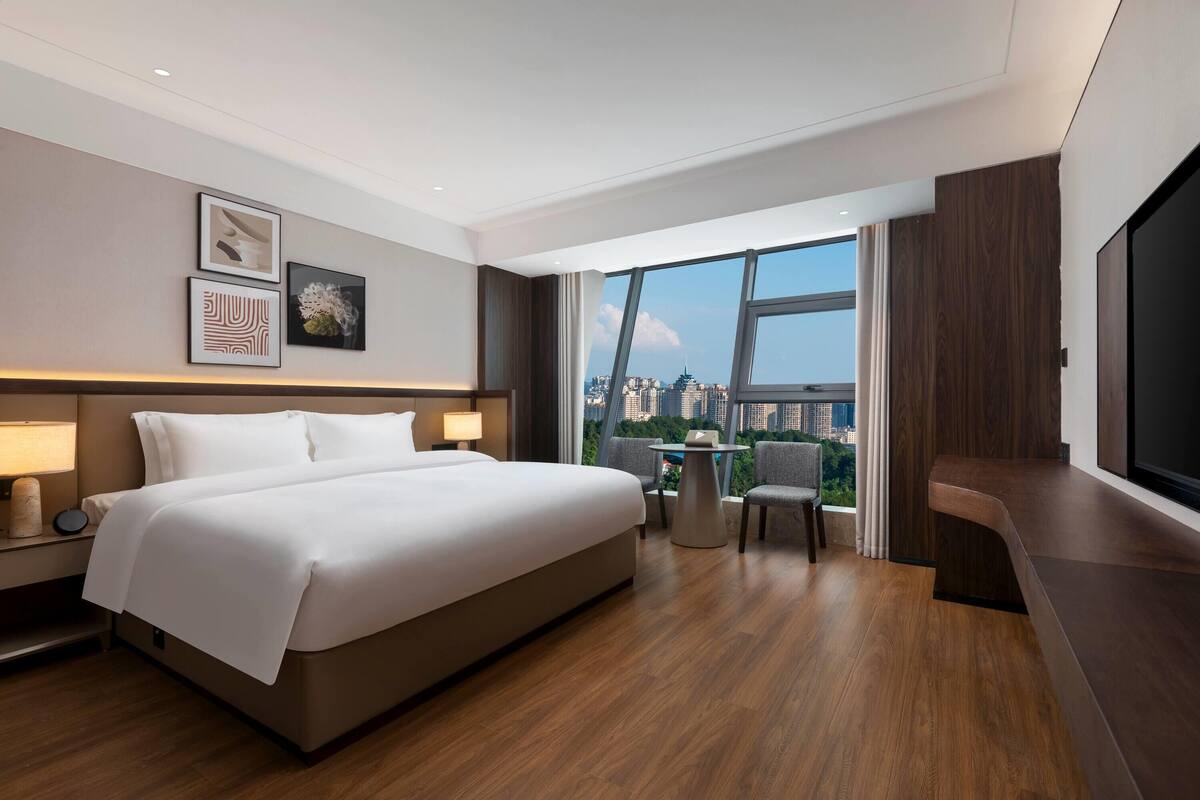 A modern hotel room with a large bed, a desk with a chair, a TV, and a city view.