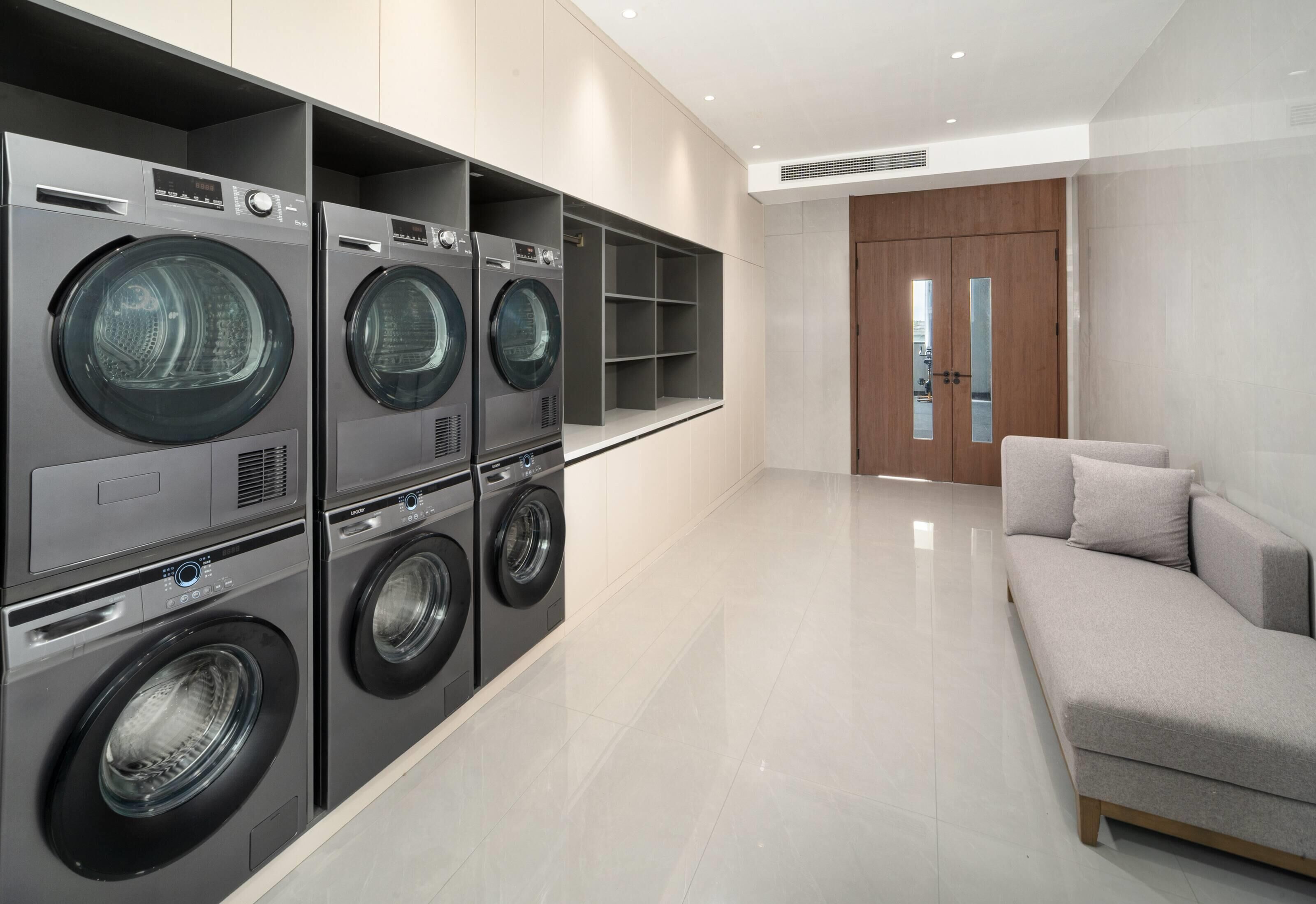 A modern laundry room with four stacked front-loading washing machines, a grey sofa, and a wooden door in the background.