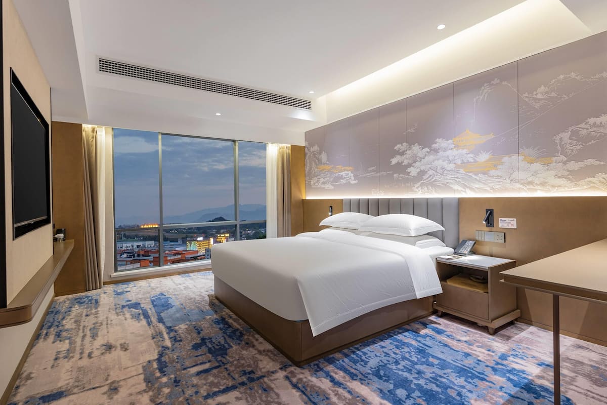 Ein modernes Hotelzimmer mit einem großen Bett, einem Schreibtisch, einem Nachttisch und einem großen Fenster mit Blick auf die Stadt.