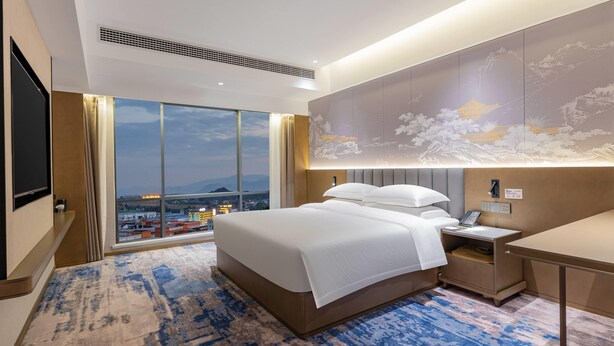 A modern hotel room with a large bed, a desk, a nightstand, and a large window offering a city view.