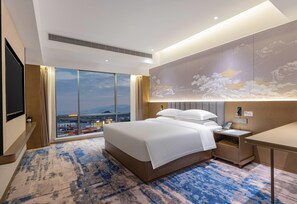 Una habitación de hotel moderna con una cama grande, un escritorio, una mesita de noche y un ventanal con vistas a la ciudad.