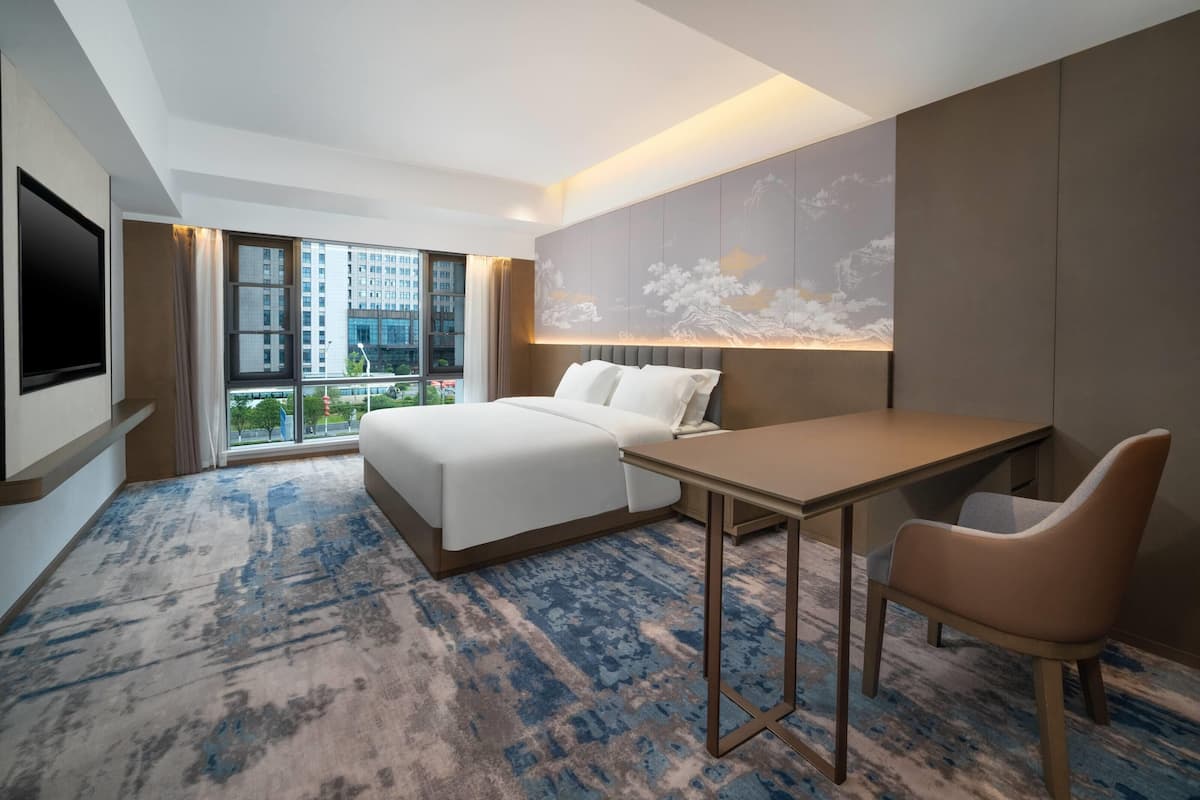 Ein modernes Hotelzimmer mit einem großen Bett, einem Schreibtisch und einem Sessel. Ein Fernseher ist an der Wand befestigt und es gibt ein Fenster mit Blick auf Gebäude.