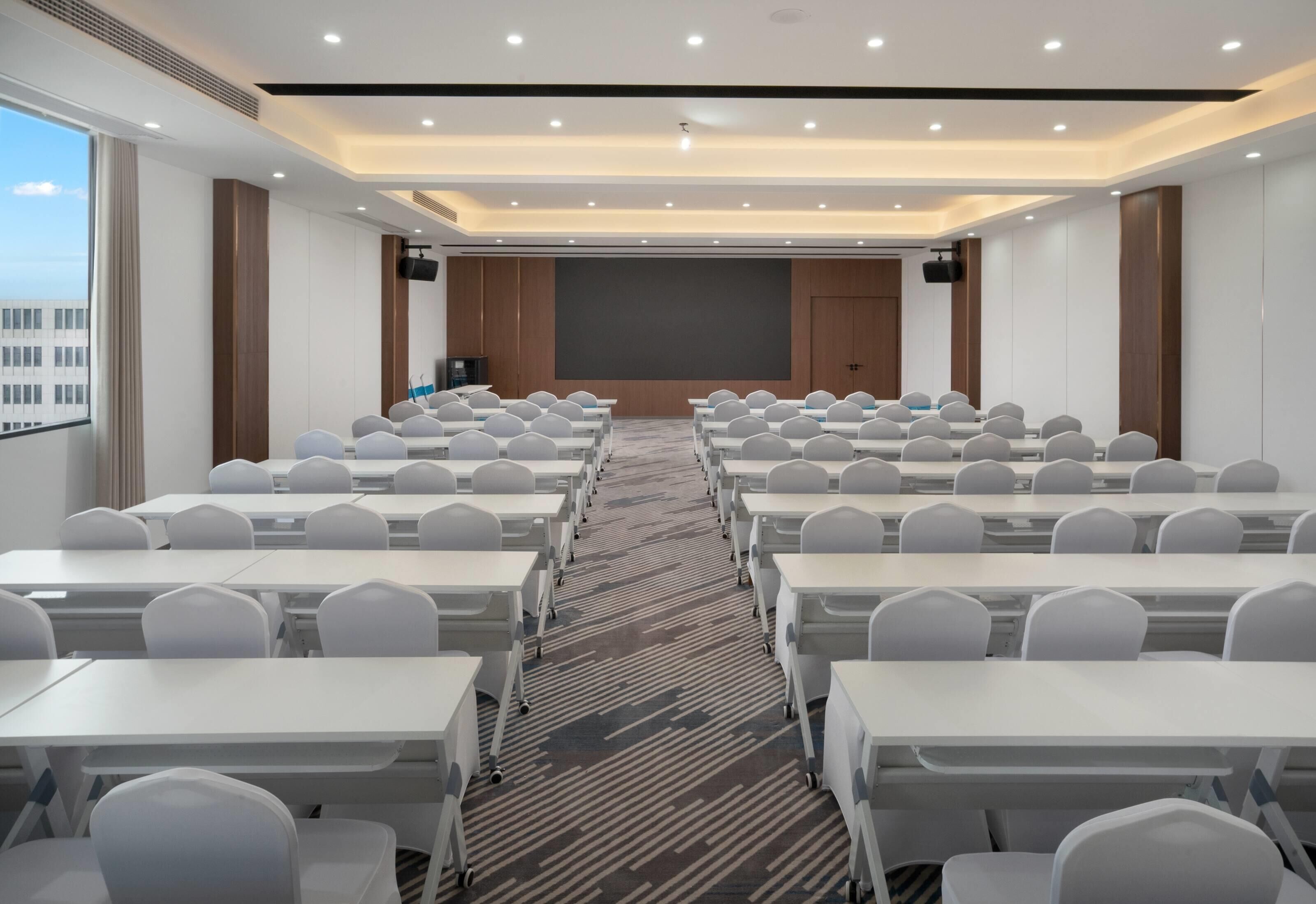 A conference room with rows of white tables and chairs, a large screen at the front, and a carpeted floor.