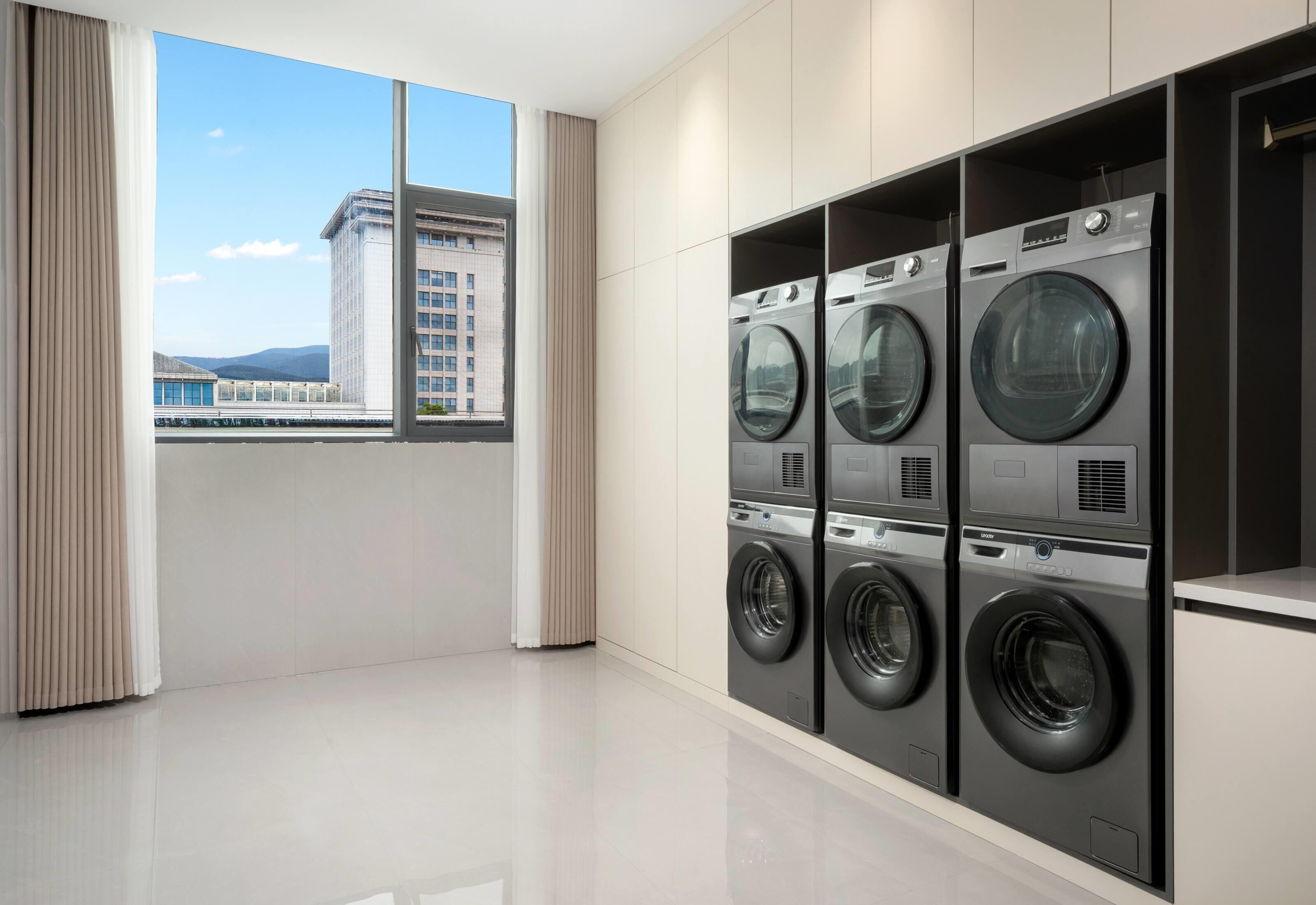 Un moderno lavadero con cuatro lavadoras de carga frontal, amplias ventanas con vista a un paisaje urbano y un diseño limpio y minimalista.