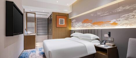 A modern hotel room with a large bed, a bedside table with a phone and a lamp, a flat-screen TV mounted on the wall, and a decorative wall panel.