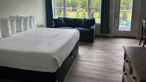 A bedroom with a bed, a sofa, a wooden dresser, and a view through a large window. - Juniper Motel-King Suite-RM12*Efficiency Kitchen*Lakefront*ATV Access (Greenville)