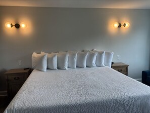 A neatly made bed with a white duvet and multiple pillows, two bedside tables, and wall-mounted lighting. - Juniper Motel-King Suite-RM12*Efficiency Kitchen*Lakefront*ATV Access (Greenville)