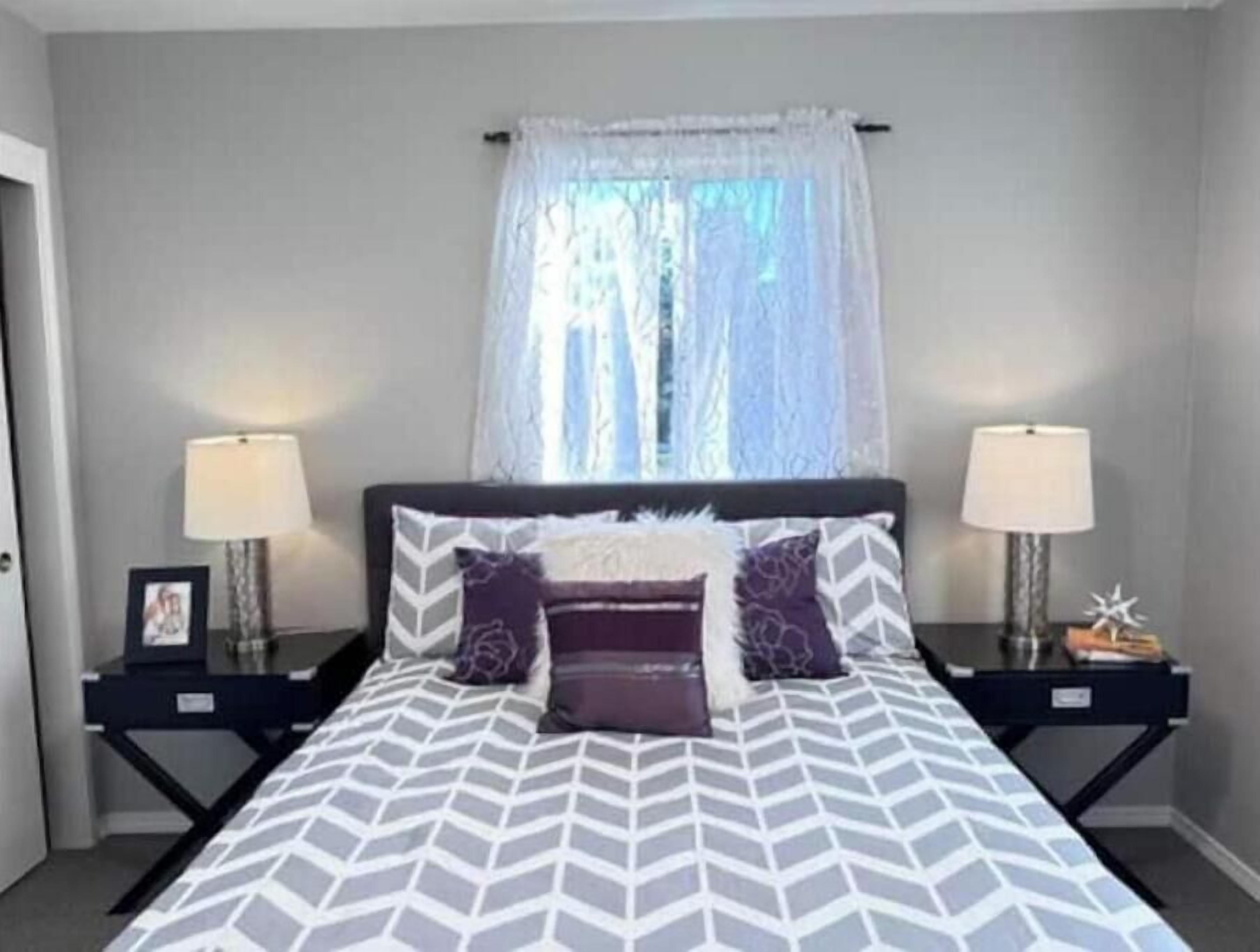 A bedroom with a bed, two bedside tables, two lamps, a window with a sheer curtain, and a picture frame on one of the tables.