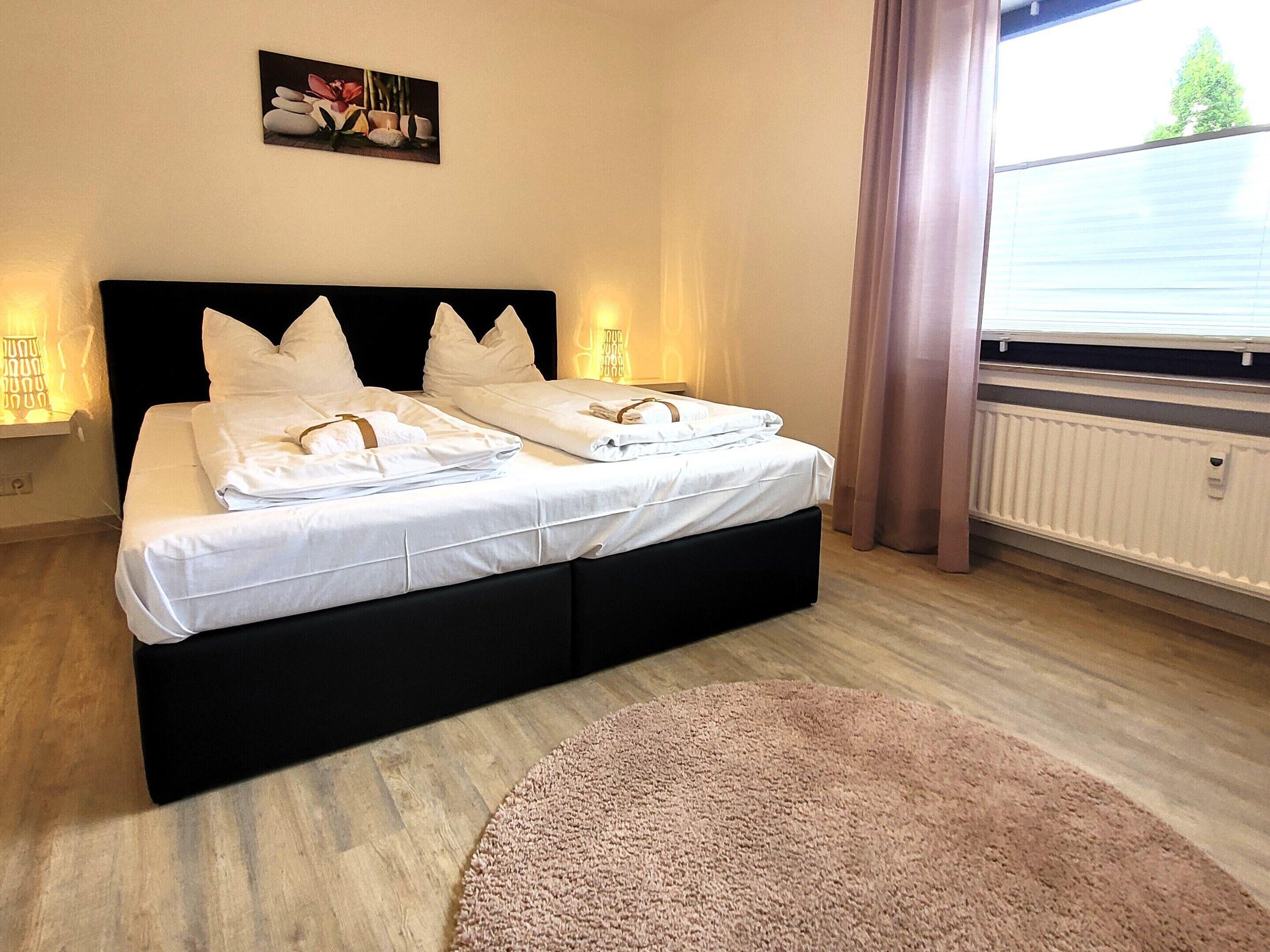 Una cama doble con ropa de cama y almohadas blancas, un cabecero negro, una alfombra beige redonda, un cuadro en la pared y una ventana con cortinas.